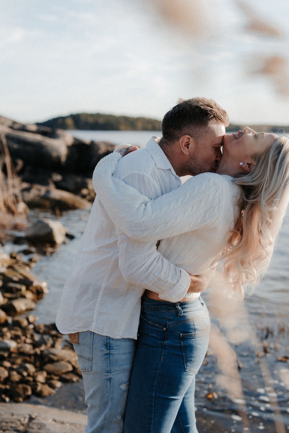 A couple shares a passionate kiss on a rocky beach, both wearing white shirts and jeans, with ocean waves in the background.