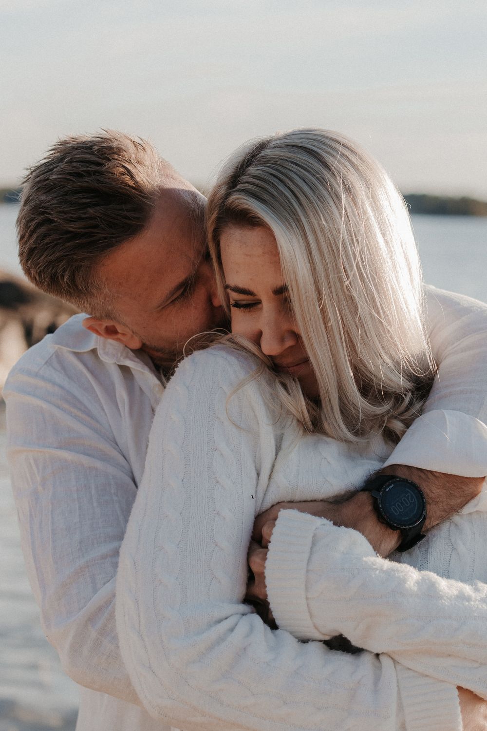 A couple in white sweaters share a tender embrace by the water at golden hour.