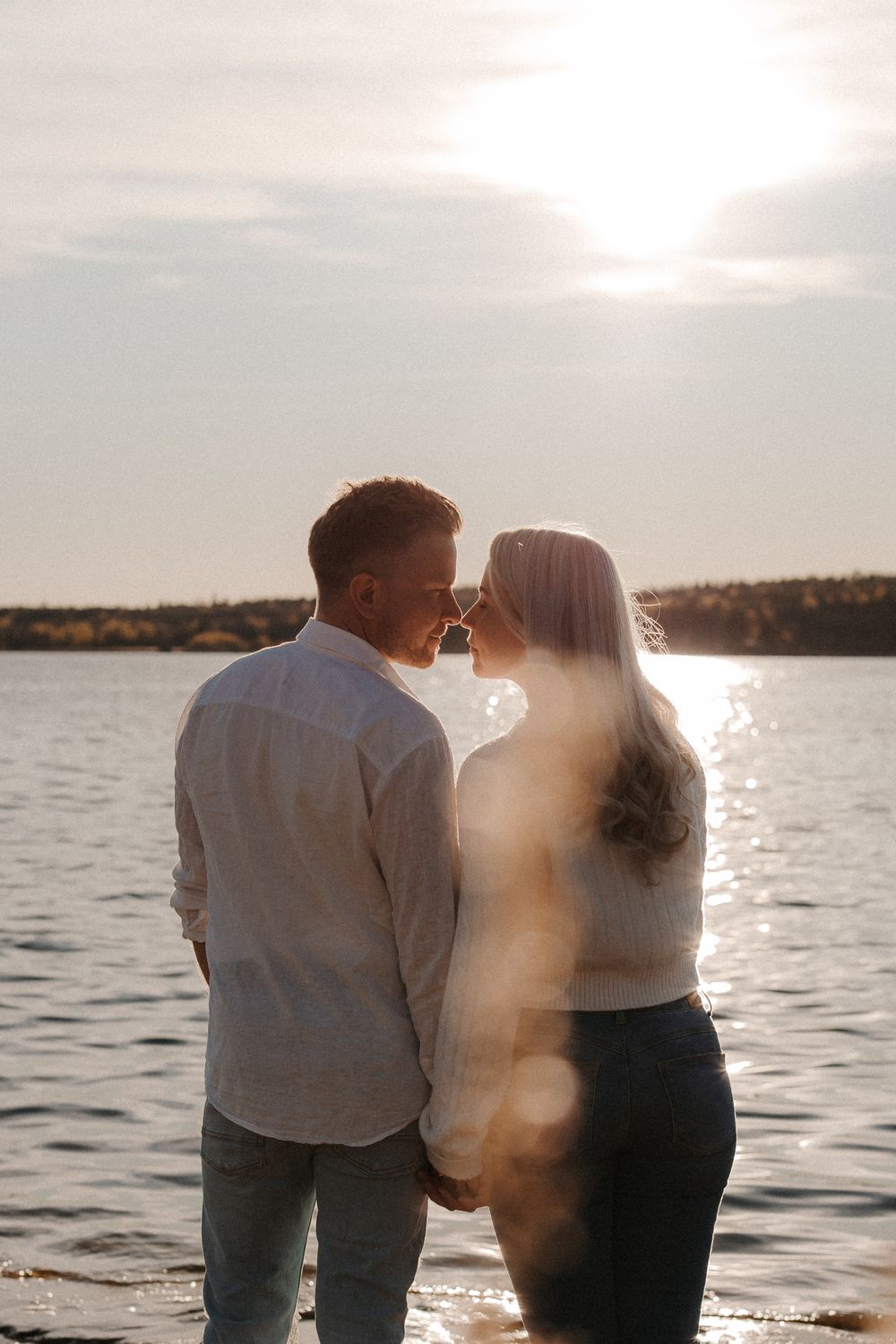 Couple sharing a romantic moment by a sunlit lake at golden hour, backlit by warm glowing sunlight.