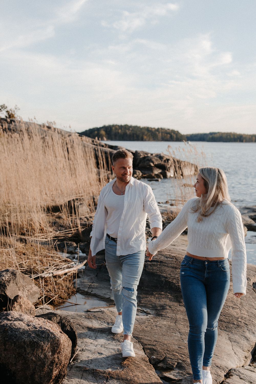 Couple holding hands and walking along rocky shoreline with tall reeds and calm water in background.