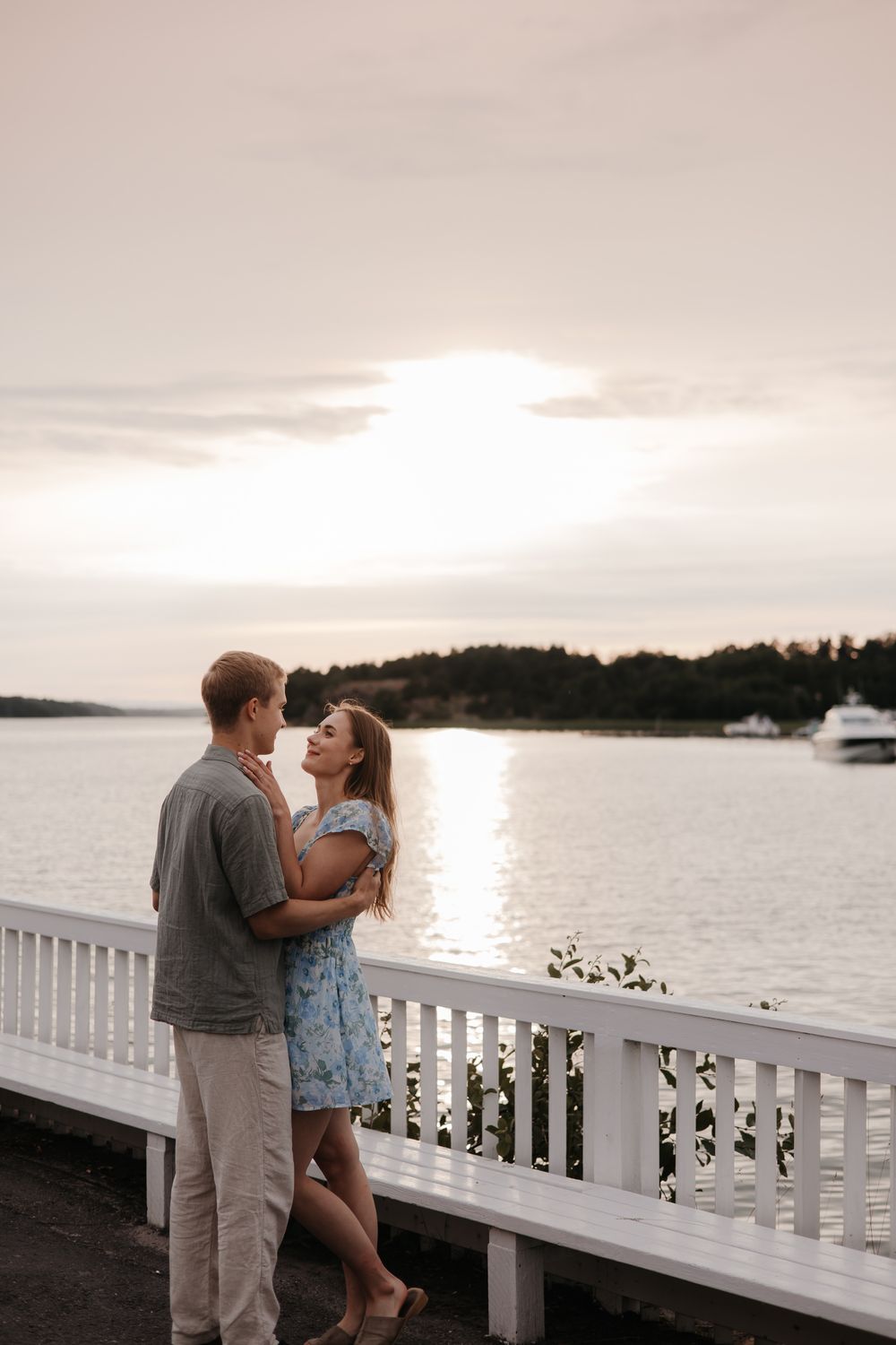 A couple shares a romantic moment on a white railing deck overlooking a serene lake at sunset.