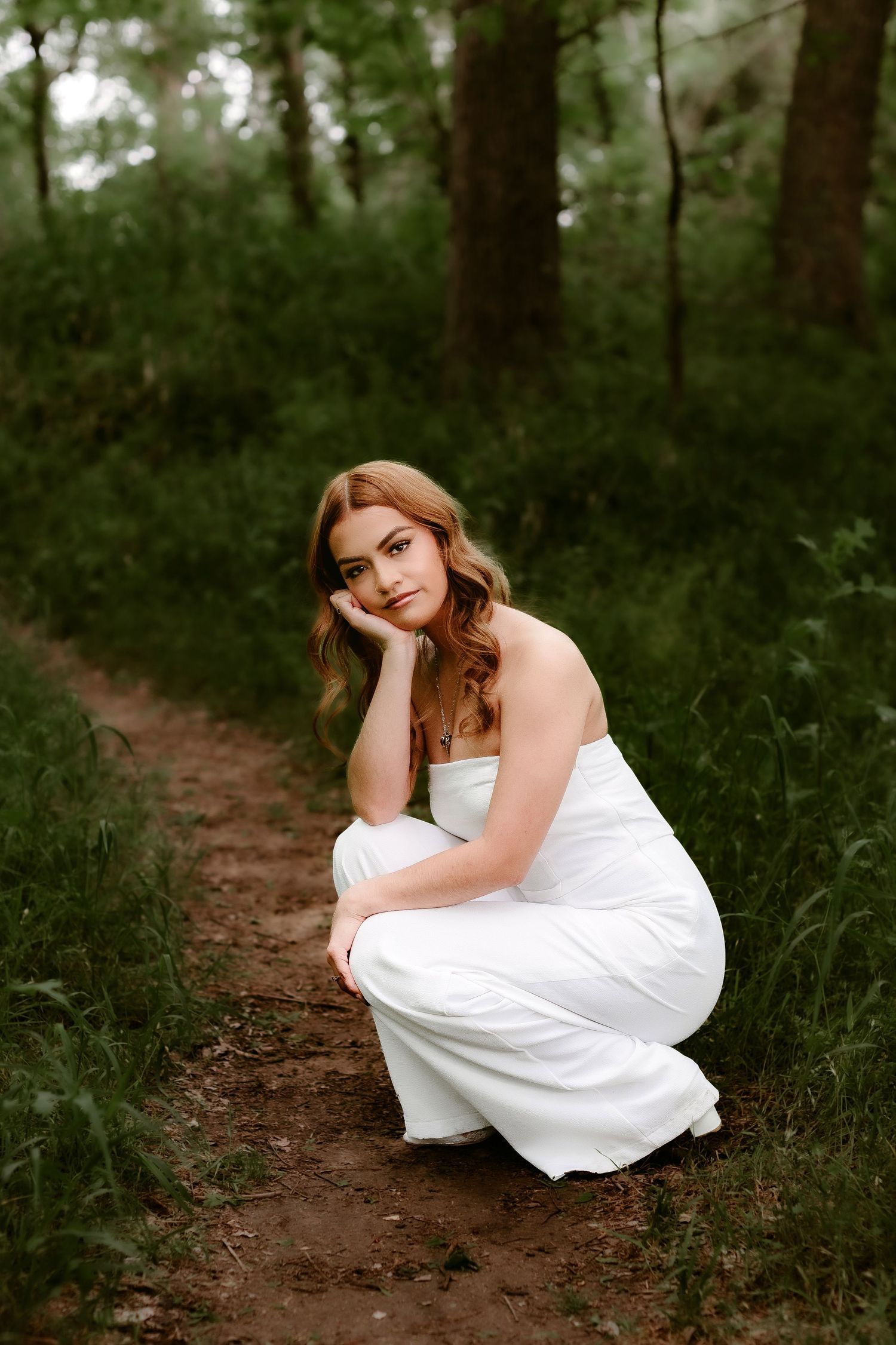 A woman in a white dress crouches on a forest path surrounded by lush greenery.