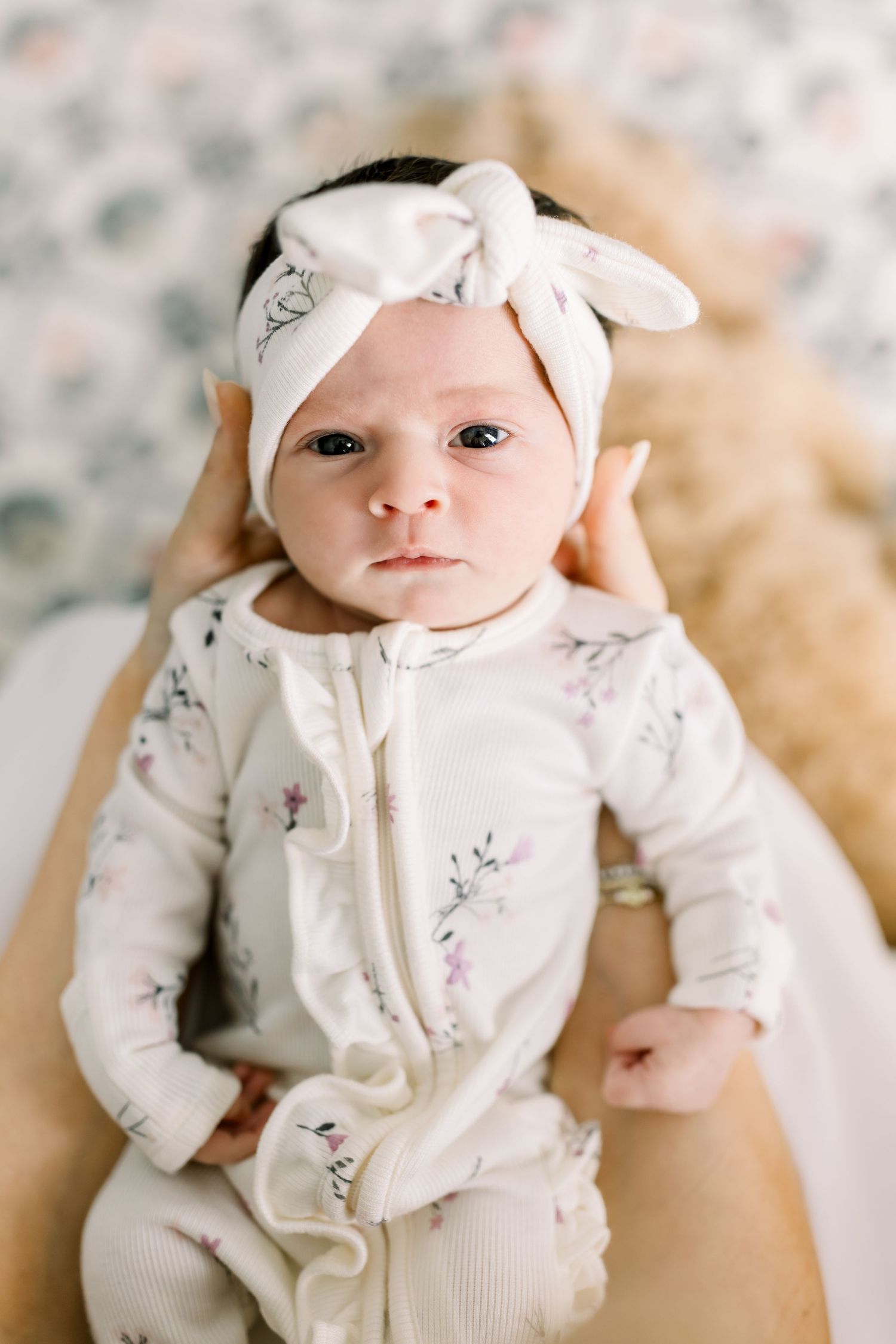 Baby in white floral outfit with bow headband looks directly at camera.