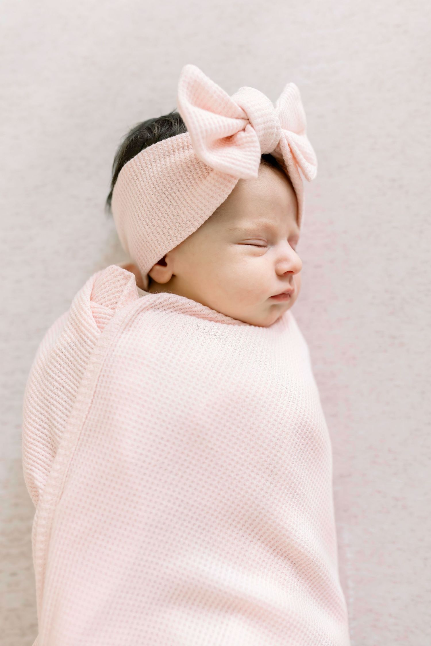 Peaceful sleeping baby wearing pink bow headband and soft swaddle.