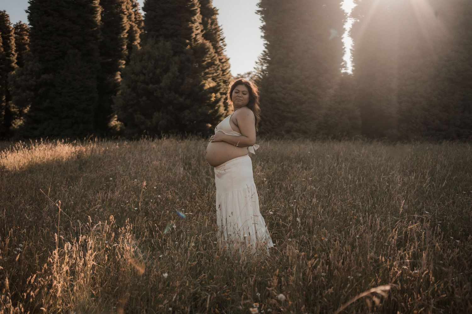 Maternity photo of expectant mother in white dress at sunset among tall trees.