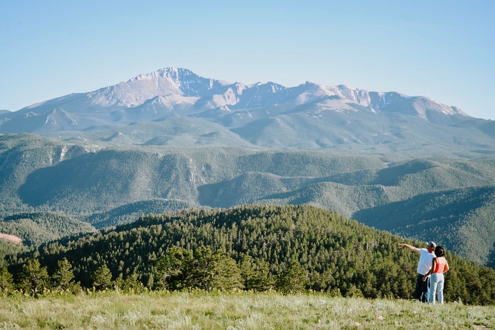 Two people stand on a grassy hill overlooking Pikes Peak and forested mountains in Colorado.