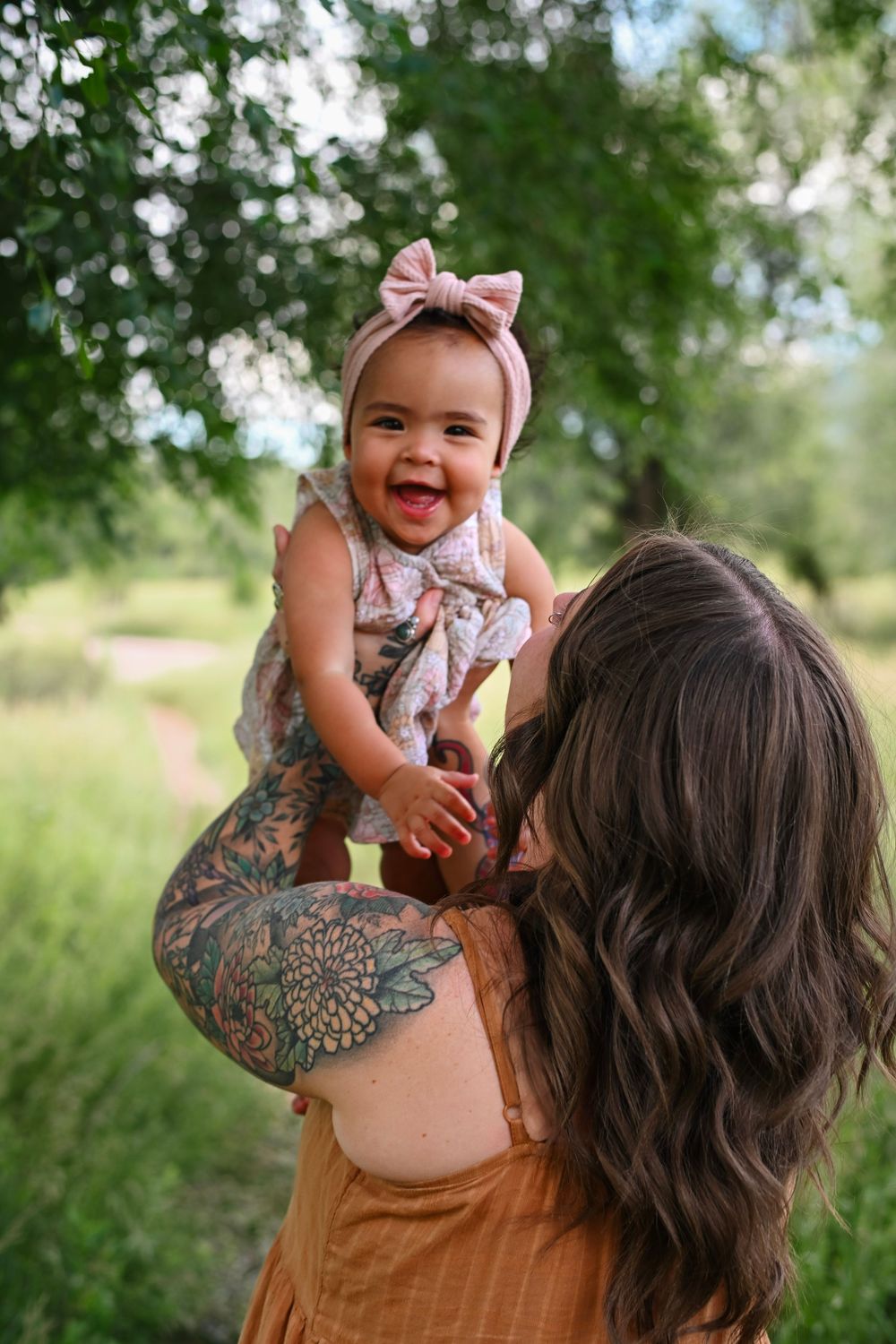 A baby wearing a pink bow headband and floral outfit is held up outdoors by someone with long brown hair and tattoos.