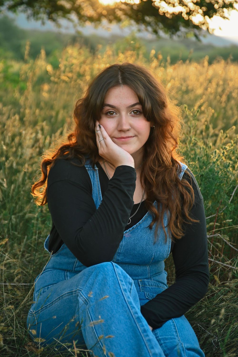 A golden sunset illuminates a field where someone poses in a denim jumpsuit and black sweater.