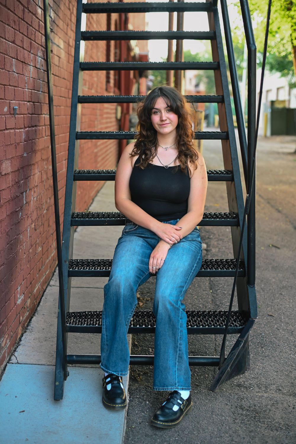 Someone in casual attire sits on black metal fire escape stairs against a brick wall backdrop in an urban setting.