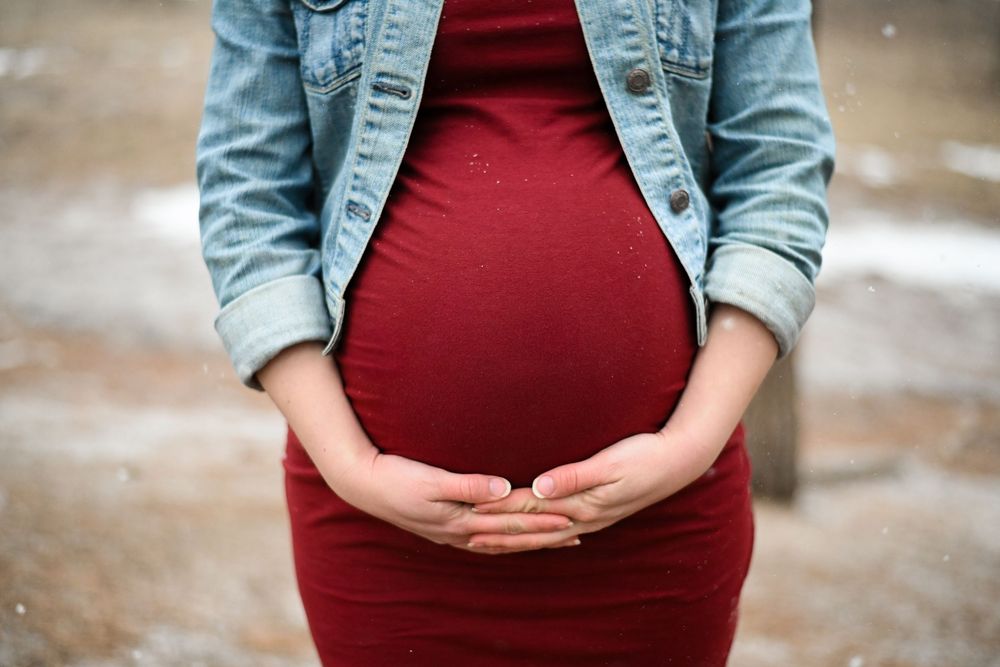 A pregnant woman in a burgundy dress and denim jacket gently cradles her baby bump against a soft blurred background.