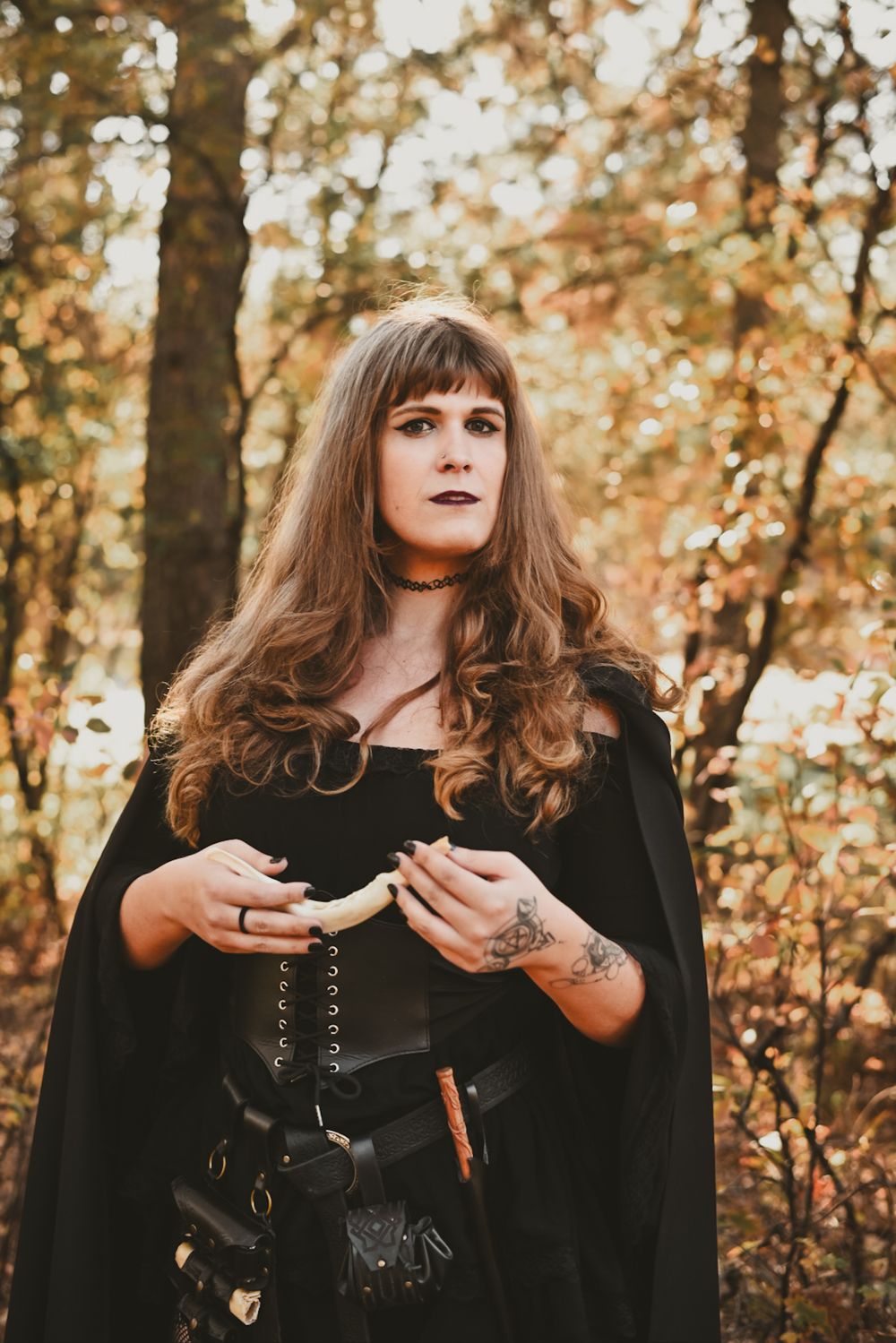 A person wearing a black cape and corset poses in the woods during autumn with golden foliage in the background.