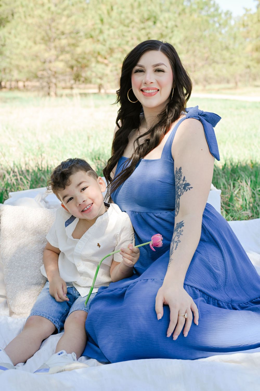 A parent and child sitting together on a blanket outdoors on sunny grass field with pine trees in background.
