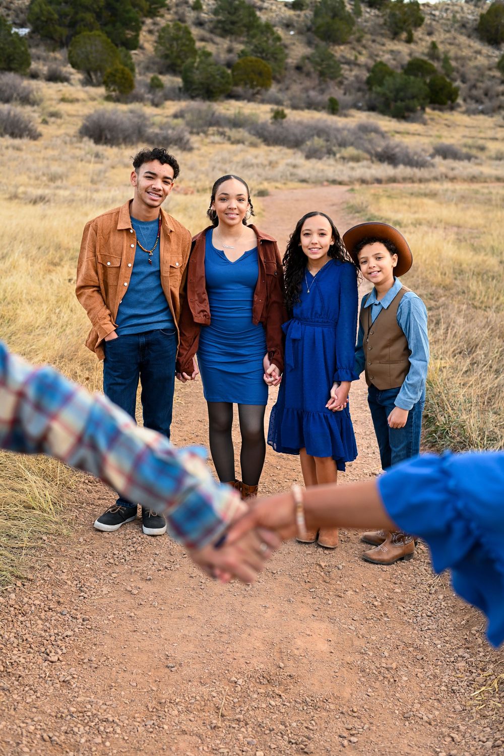 Group of people wearing coordinating blue outfits posing on a dirt path with someone's outstretched hand in foreground.