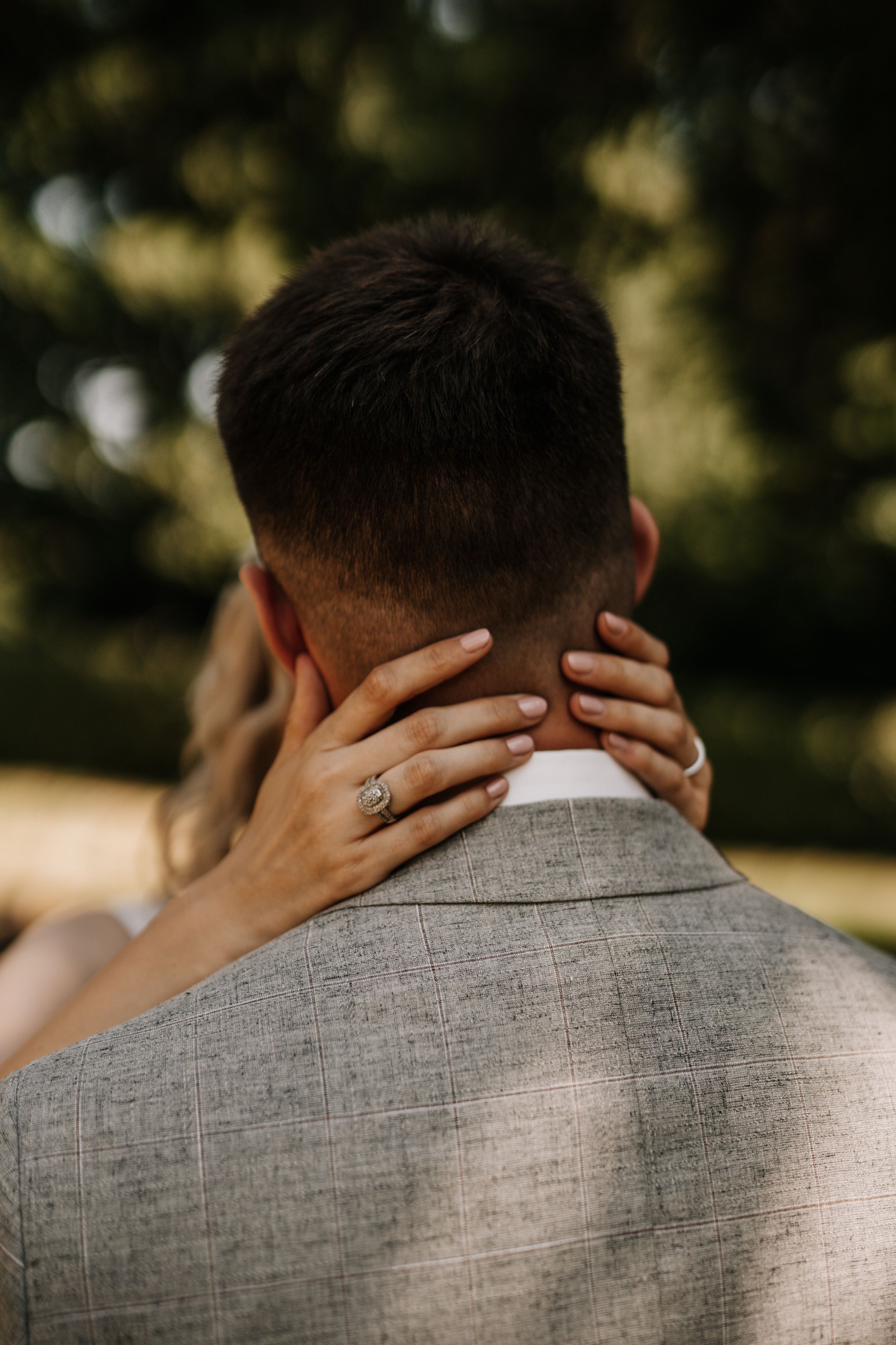Hands with engagement ring embracing groom's neck in outdoor setting.