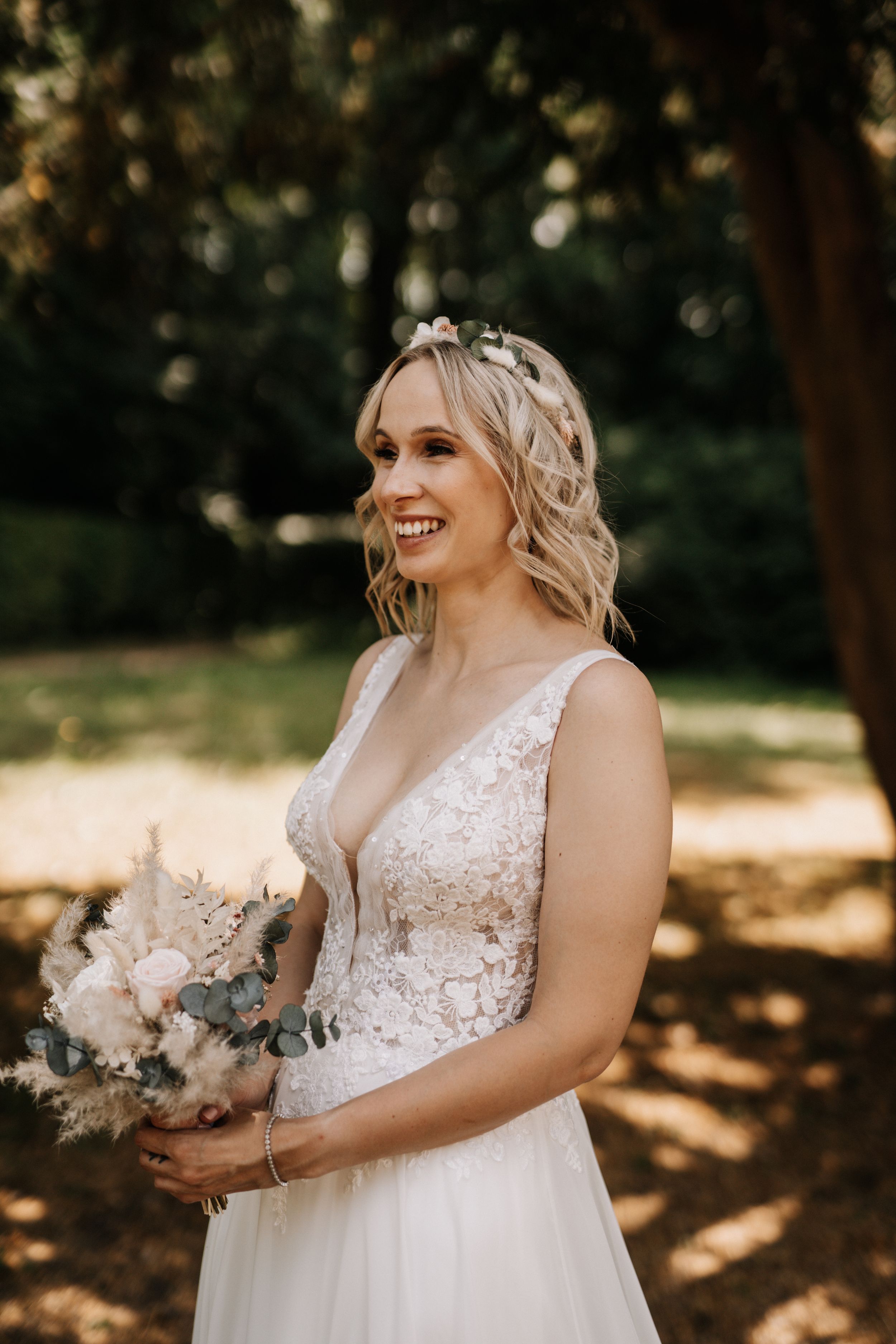Bride holding a delicate bouquet smiles joyfully in natural outdoor lighting.