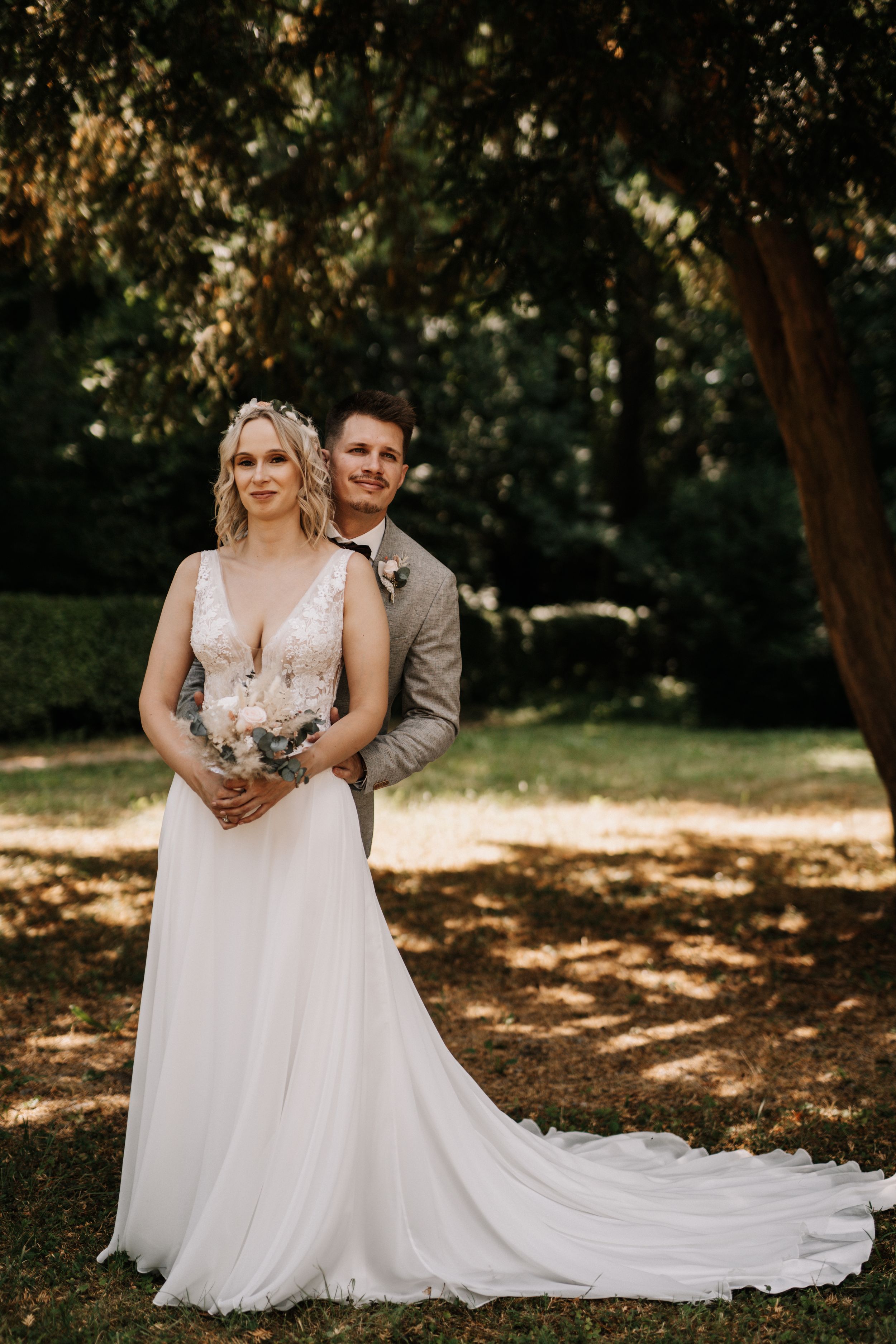 Wedding couple poses together in a wooded setting with dappled sunlight.