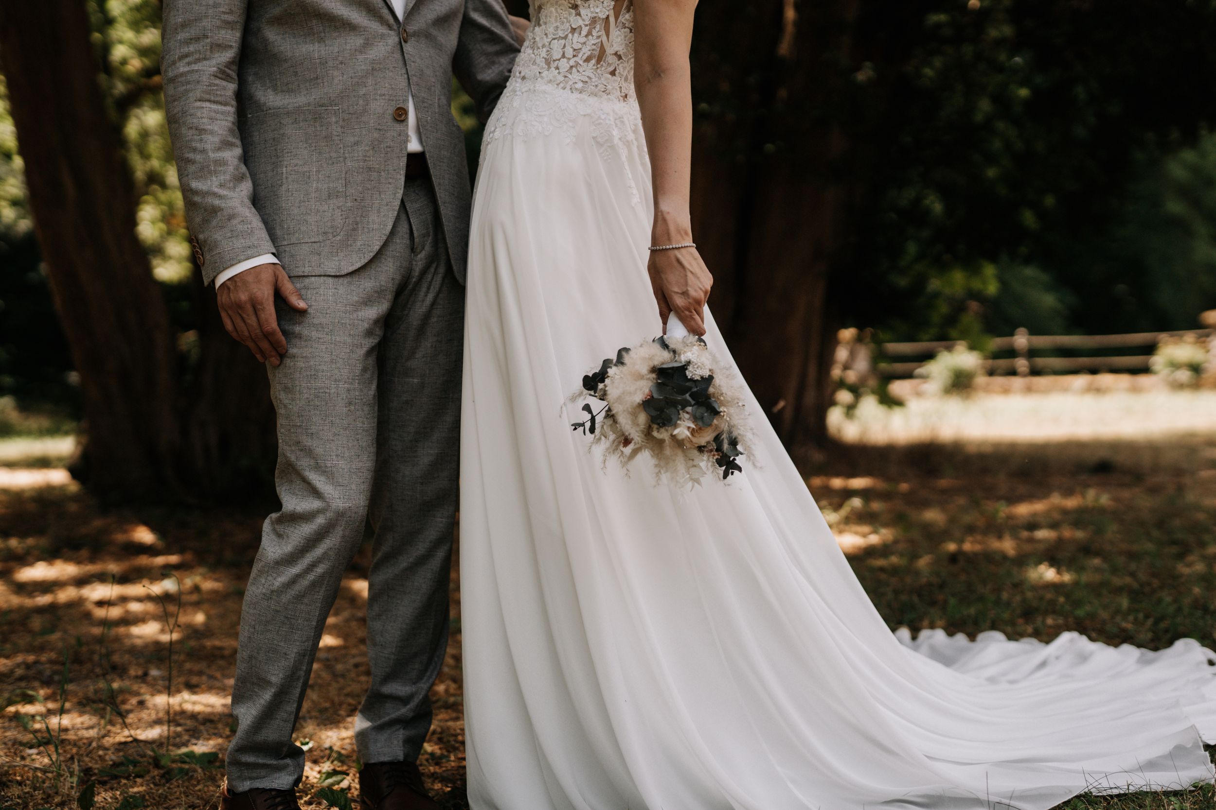 Bride in lace gown and groom in grey suit pose in wooded outdoor setting.