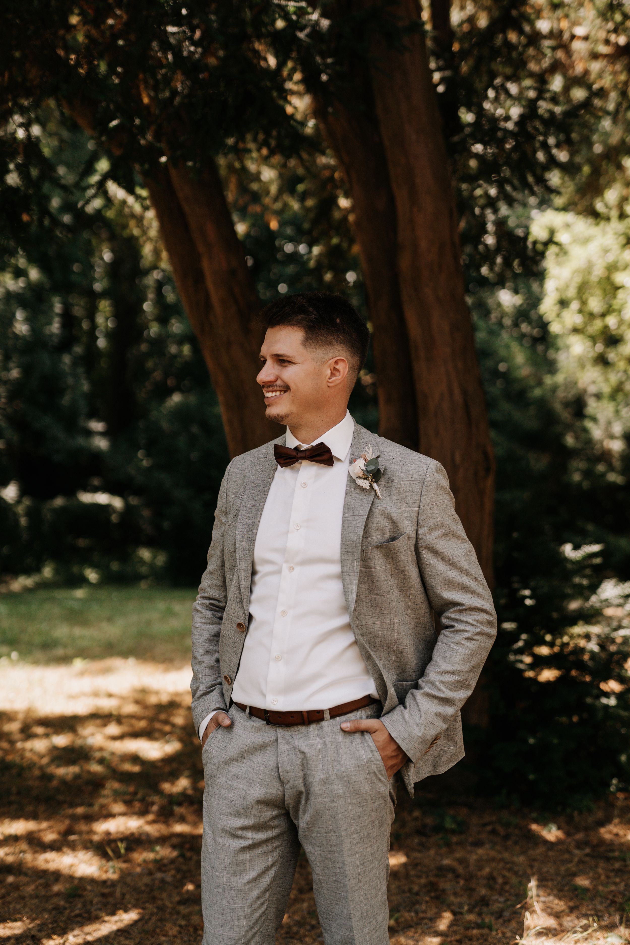 Groom in grey suit poses casually against natural wooded backdrop.