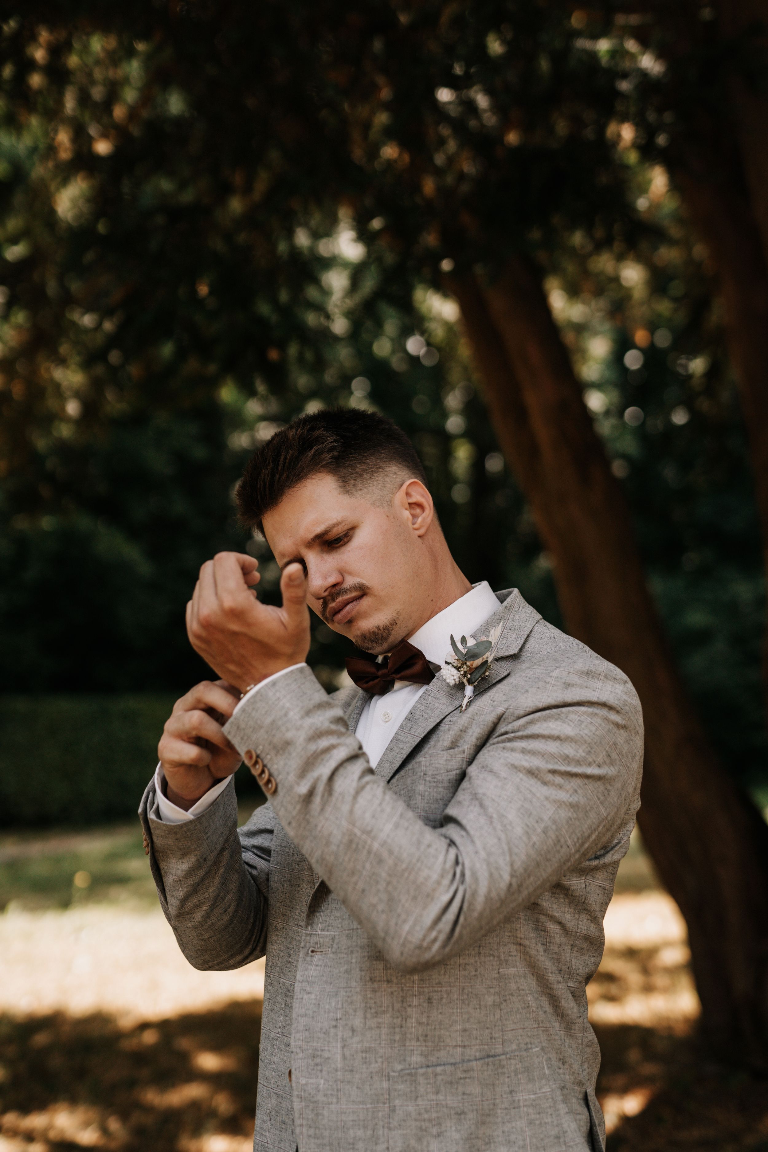 A groom in a light gray suit and bowtie wipes tears from his eyes in an emotional outdoor moment.