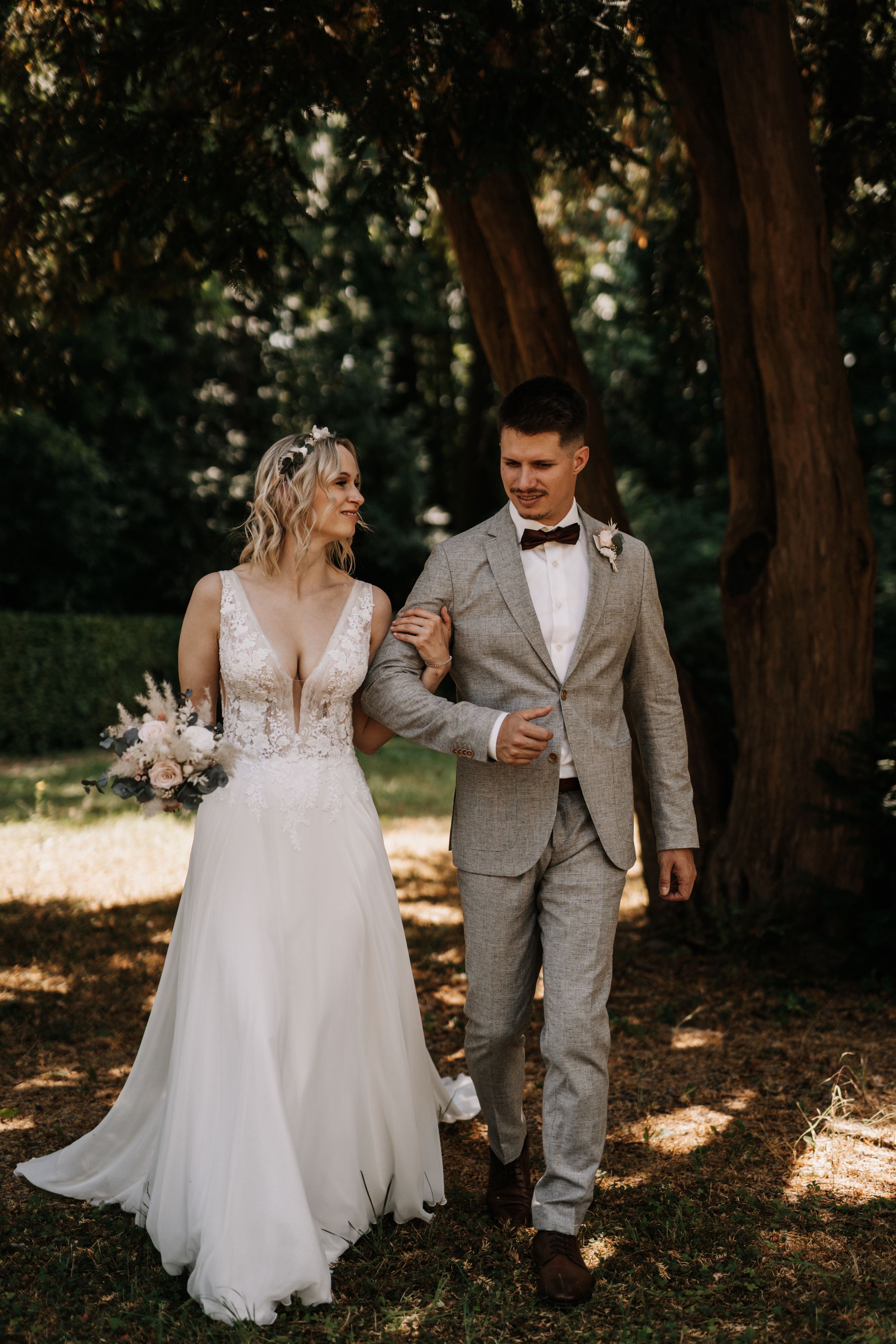 A couple walks together in a wooded setting, bride in flowing white gown and groom in light gray suit.