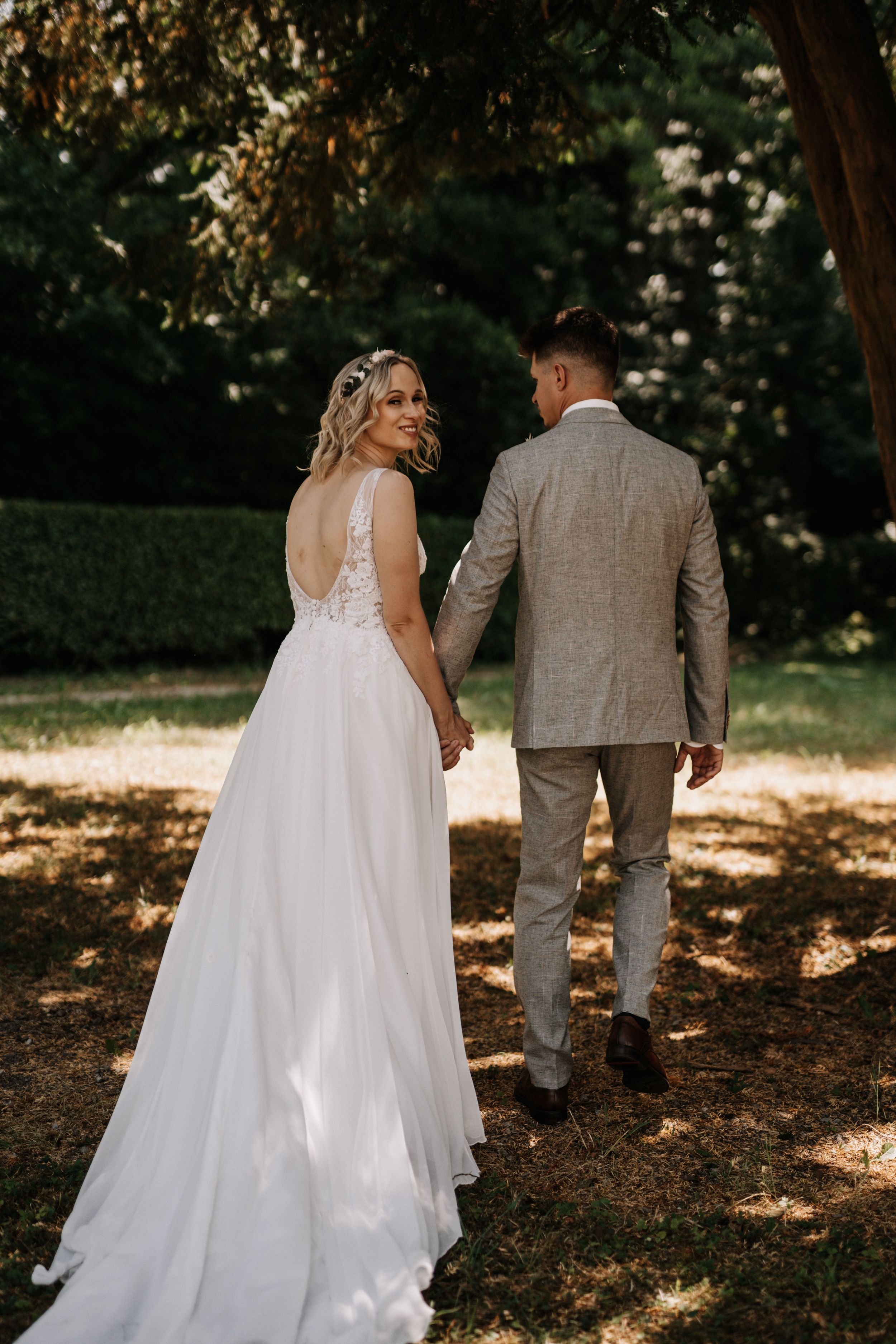 A couple walks hand in hand through a sun-dappled garden setting on their wedding day.