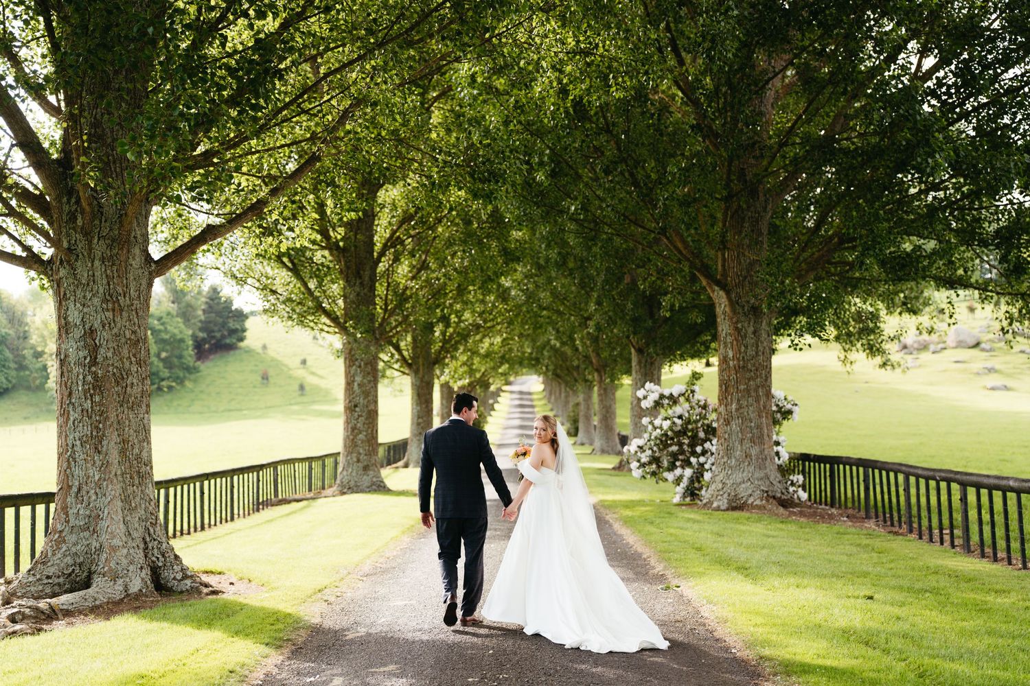 Wedding couple walks down a tree-lined path surrounded by lush greenery on a sunny afternoon.