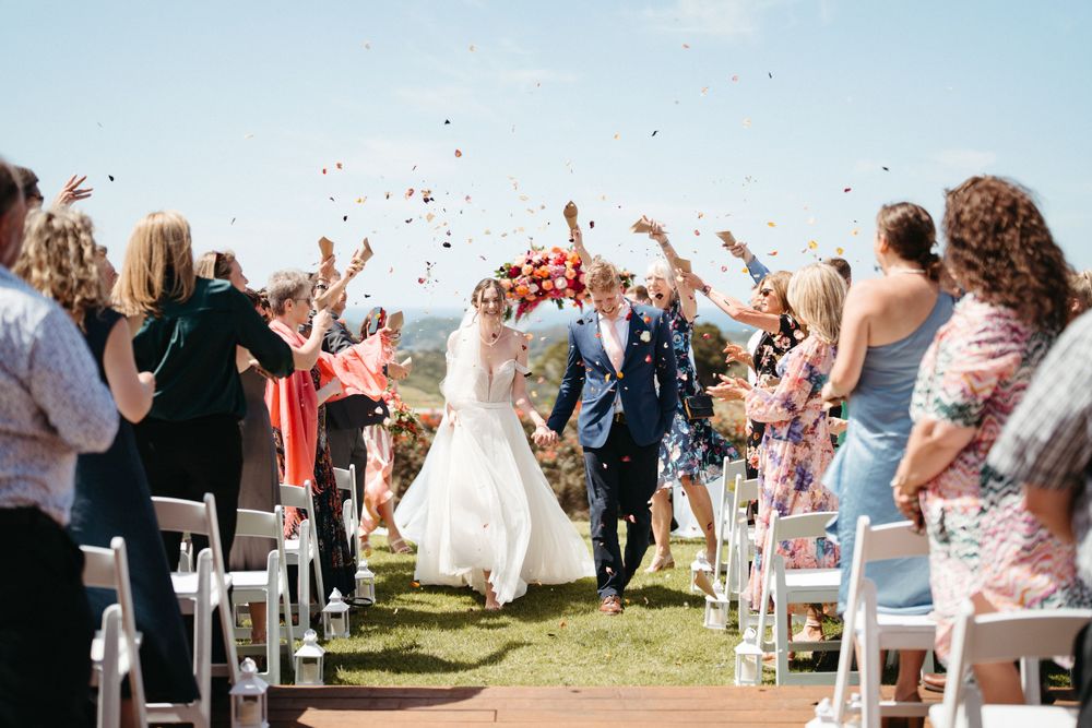 Wedding guests toss rose petals as newlyweds exit outdoor ceremony under blue sky.
