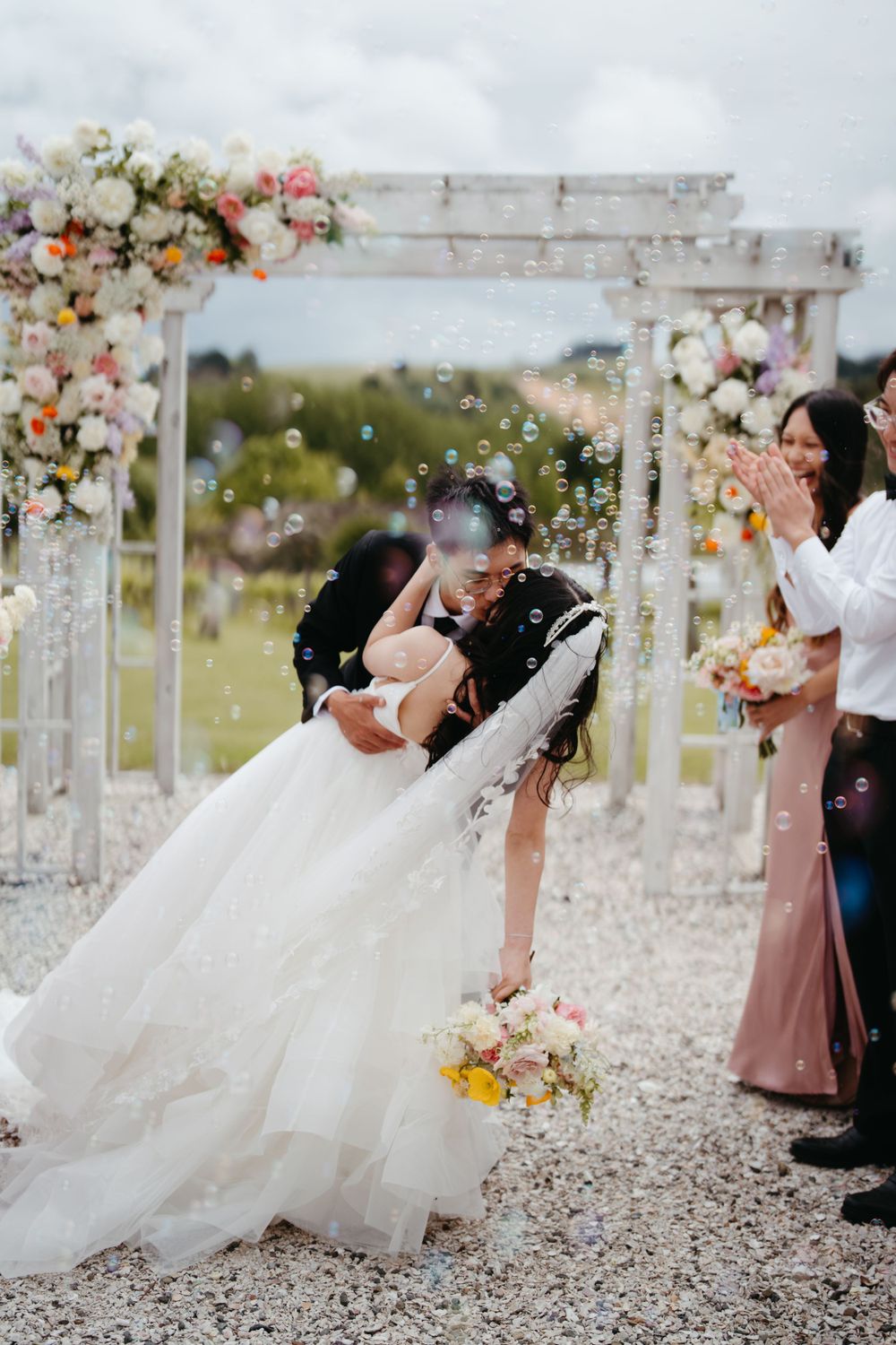 A romantic dip kiss under a floral arch with bubbles floating in the air at Turanga creek wedding venue in Auckland.