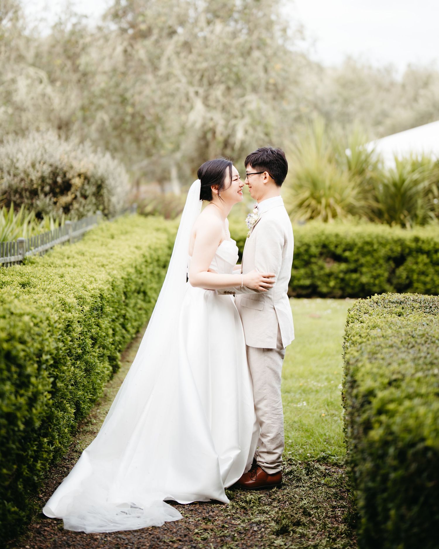 A romantic wedding couple shares an intimate moment on a bench surrounded by lush greenery in an outdoor garden setting.