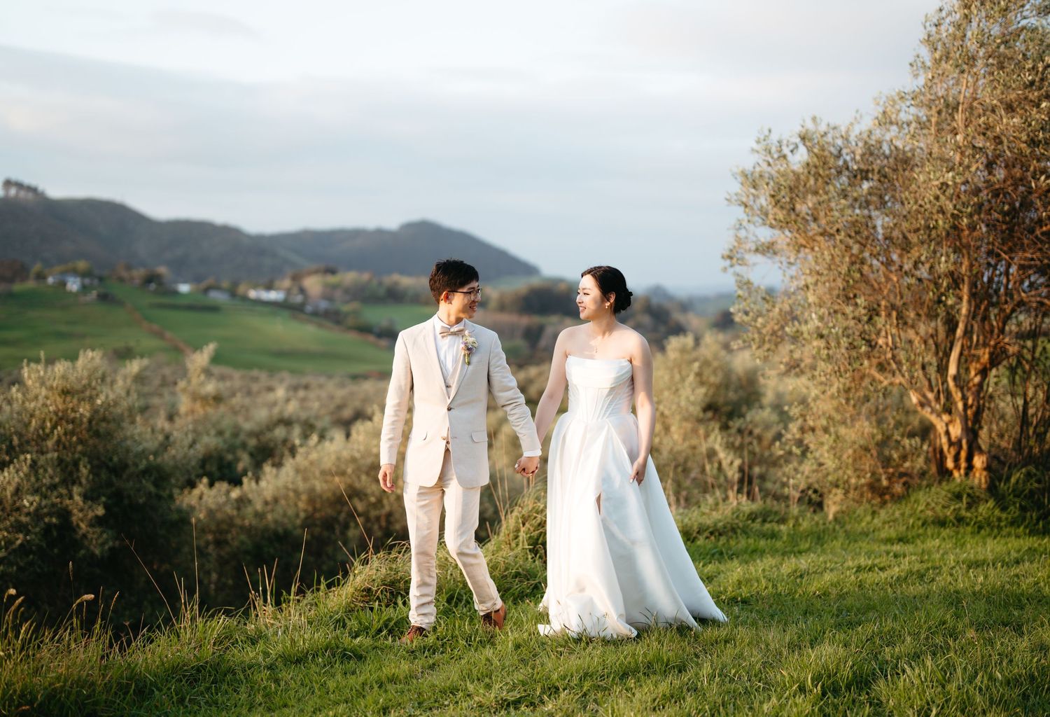 A couple in white wedding attire walks along a scenic hilltop meadow with mountains in the background during sunset.