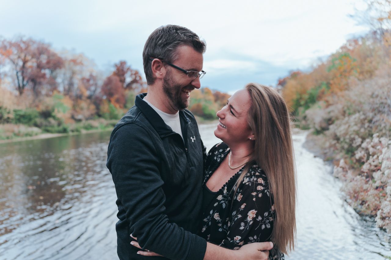 A romantic couple embracing by a river during autumn with fall foliage in the background. Summit Metro Park