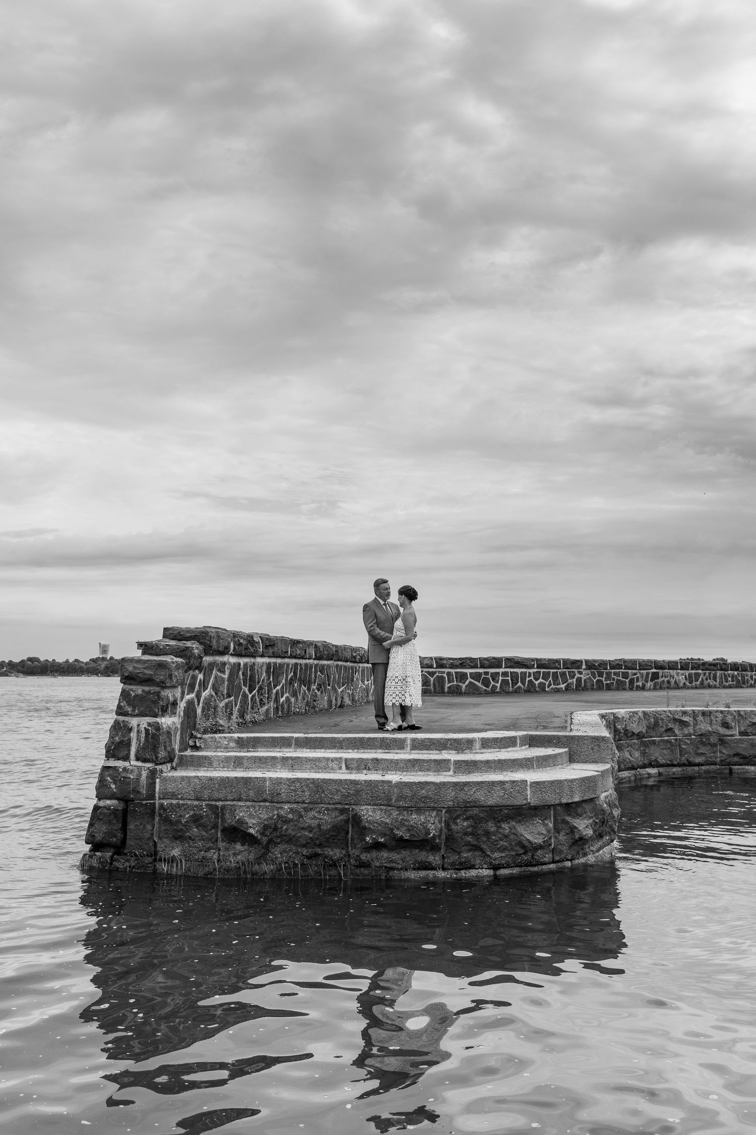 A couple stands on a stone pier overlooking calm waters on an overcast day in black and white photography.