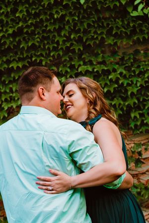 Romantic couple embraces and laughs against ivy-covered wall in coordinated mint and teal outfits.