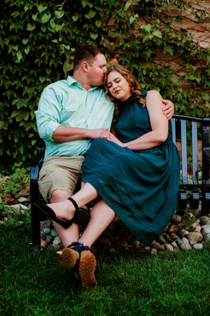 A couple shares an intimate moment while sitting together on a garden bench surrounded by green ivy and grass.