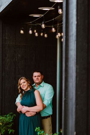 Couple embraces under romantic string lights against a dark wall during evening photoshoot.