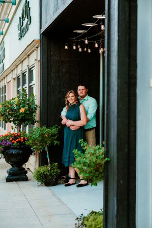 A couple stands embracing in a doorway surrounded by potted plants and flowers along a city sidewalk.