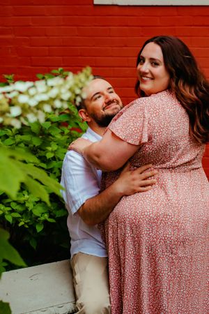 A couple embraces against a bright red wall with green foliage, wearing white and pink floral clothing.
