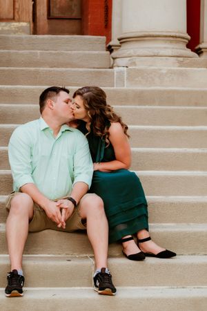 A couple in dressy casual attire shares an intimate moment while sitting on stone steps near classic columns.