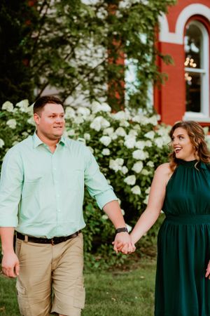 A couple in coordinating green outfits holding hands while walking near white flowering bushes and a red brick building.