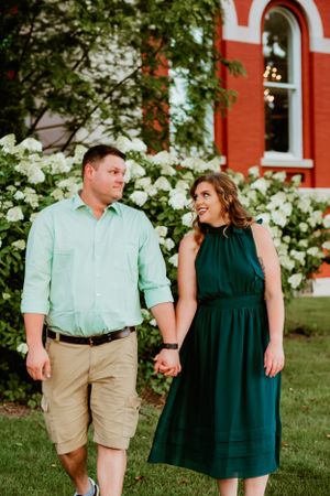 A couple holds hands while standing on grass near a red brick building with white hydrangea flowers blooming.