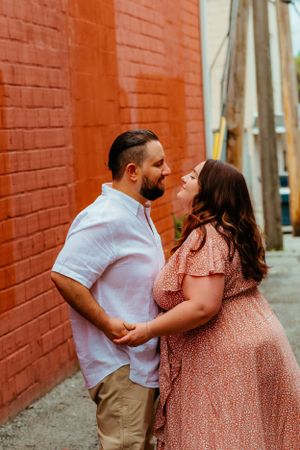 A couple shares an intimate moment in an alley with a vibrant orange brick wall backdrop during an engagement photoshoot.