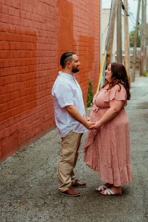 A couple holds hands in an alley, with a brick wall backdrop, as they share an intimate moment together.