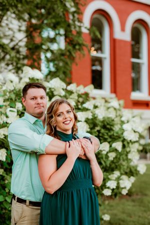 A couple poses together against a red brick building with white flowers and arched windows in the background.