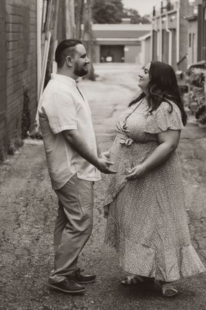A couple shares a tender moment in black and white as they stand together in an alley wearing casual summer attire.