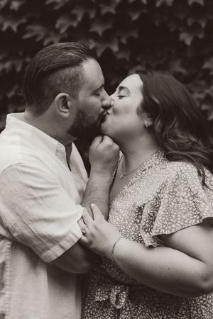 A romantic black and white portrait of a couple sharing an intimate kiss against an ivy-covered wall background.