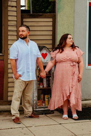 A couple stands outside a building holding hands, with her wearing a pink floral dress and him in a light blue shirt.