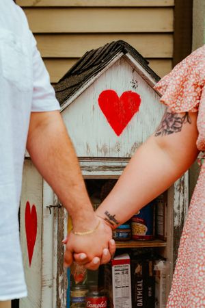 A couple holds hands in front of a rustic wooden cabinet decorated with red painted hearts.