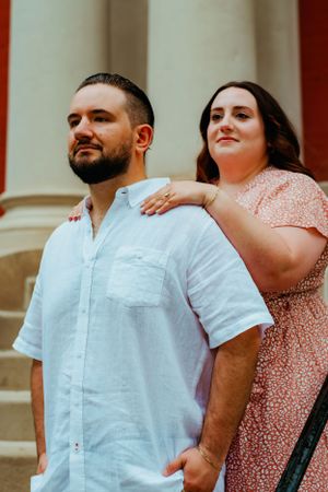 A romantic couple poses together on stone steps, with her in a pink dress and him wearing a white button-down shirt.