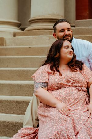 A couple sits close together on stone steps, with her wearing a pink dress and him in light blue attire.