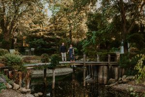 A couple stands on a wooden bridge over a tranquil pond in a lush garden during golden hour.